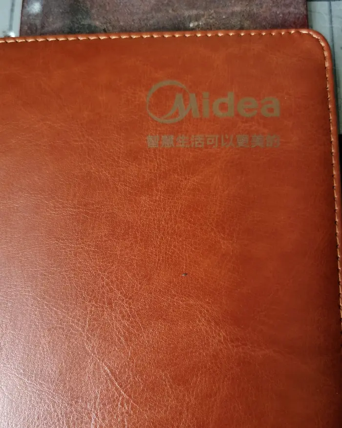 合肥激光打标代工实拍 · 棕色皮革笔记本封面激光雕刻企业 logo 与品牌标语，深浅可控批量一致